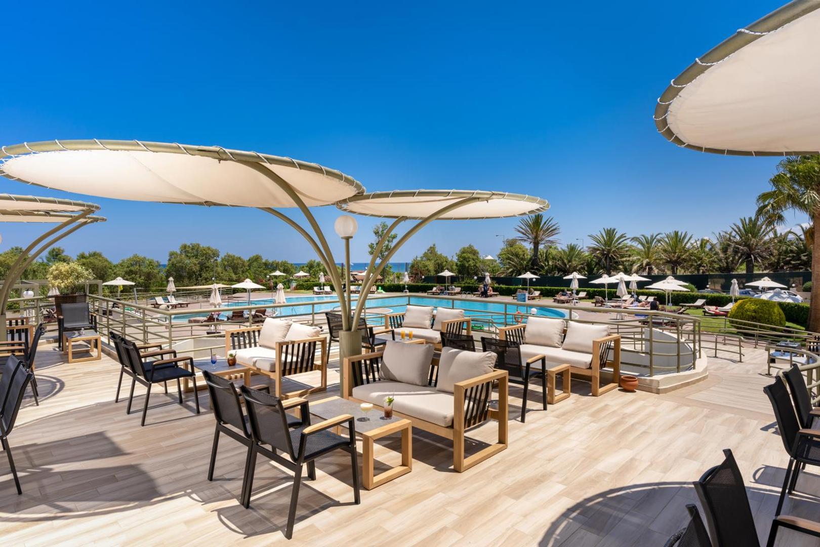 Georgioupolis Resort āra terase ar modernām mēbelēm un saulessargiem pie baseina — atpūtas zona ar skatu uz palmu dārzu