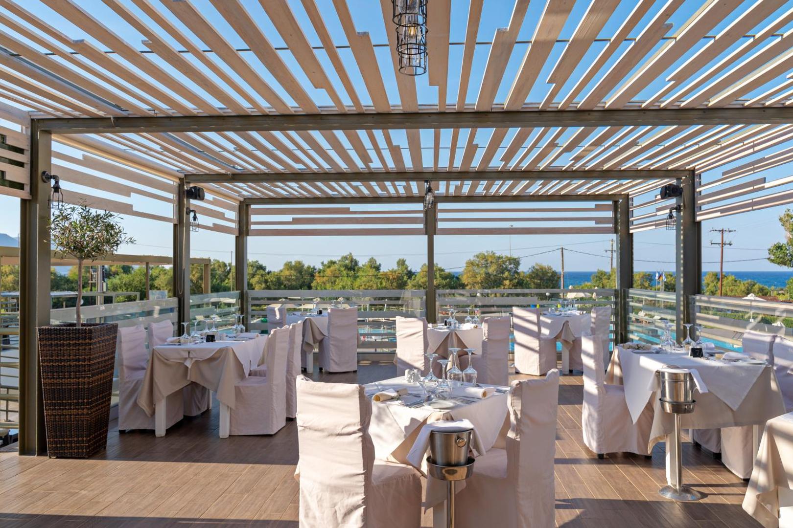 Georgioupolis Resort āra terases restorāns ar eleganti klātiem galdiem zem koka pergolas un skatu uz jūru un kūrorta dārzu
