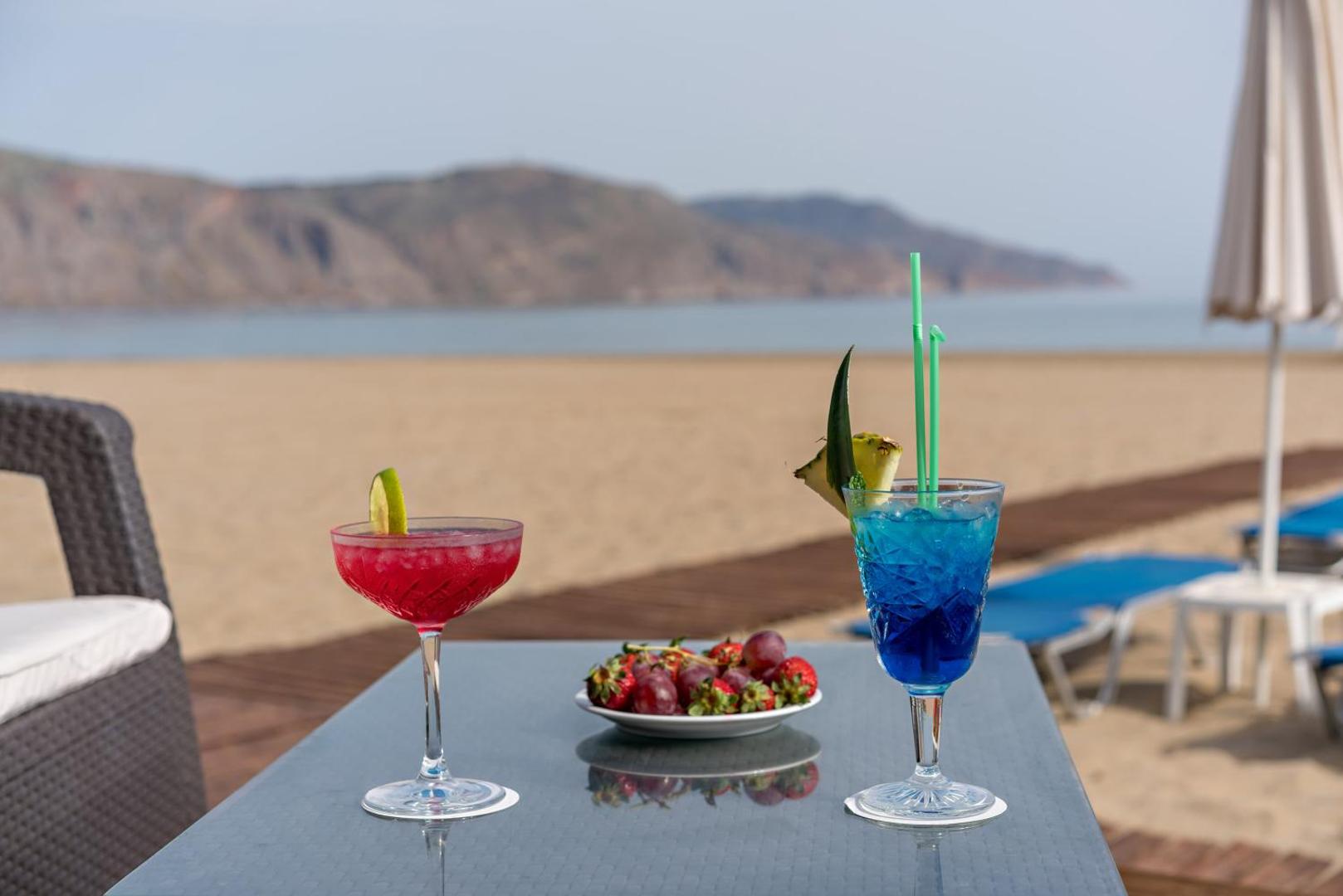 Georgioupolis Resort Aqua Park and SPA krāsaini kokteiļi un augļi uz galda pludmalē — skats uz smilšu krastu un kalniem