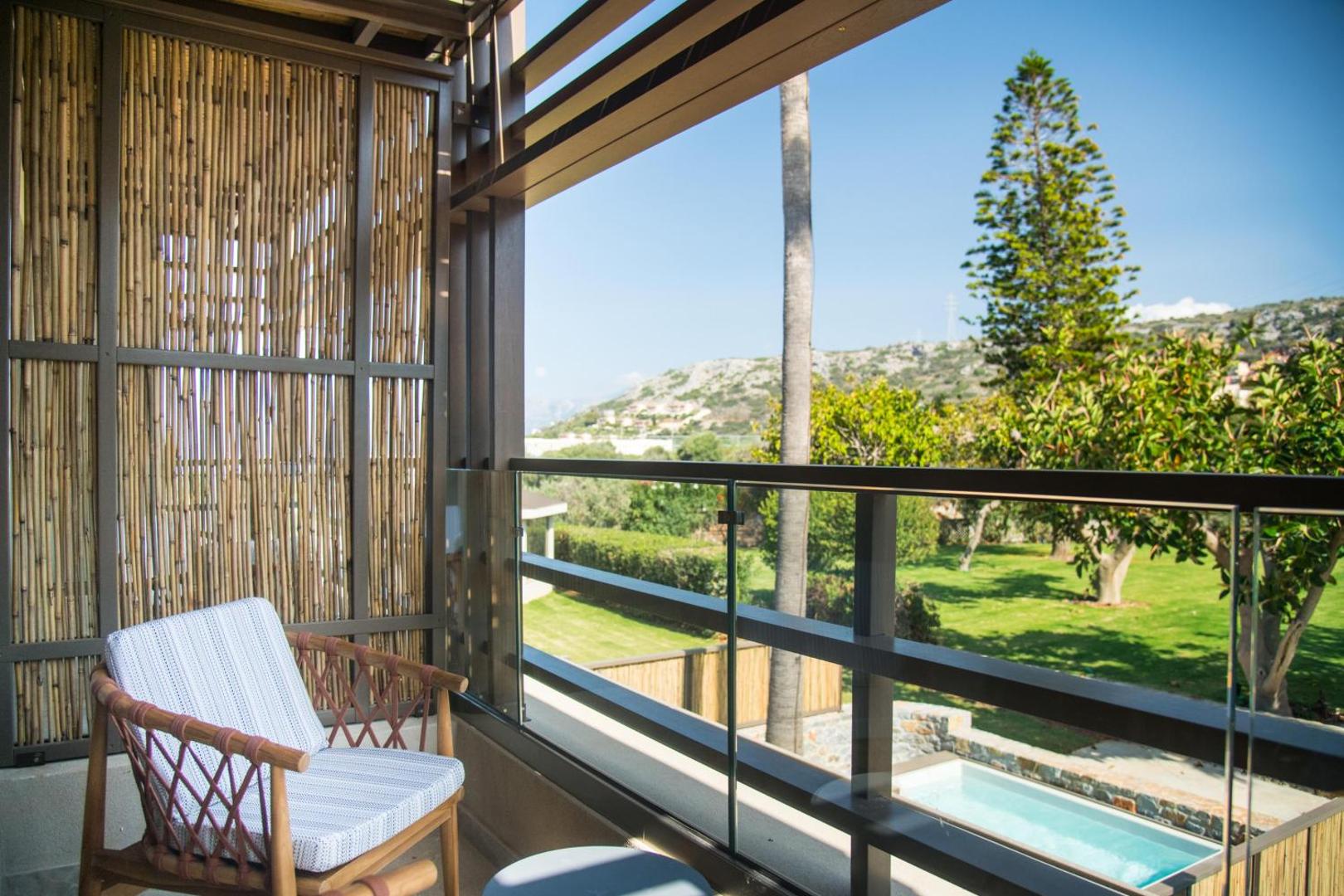 Skats no King Minos Retreat Resort & Spa numura balkona uz dārzu un nelielu baseinu — koka apdare un mājīgs atpūtas krēsls
