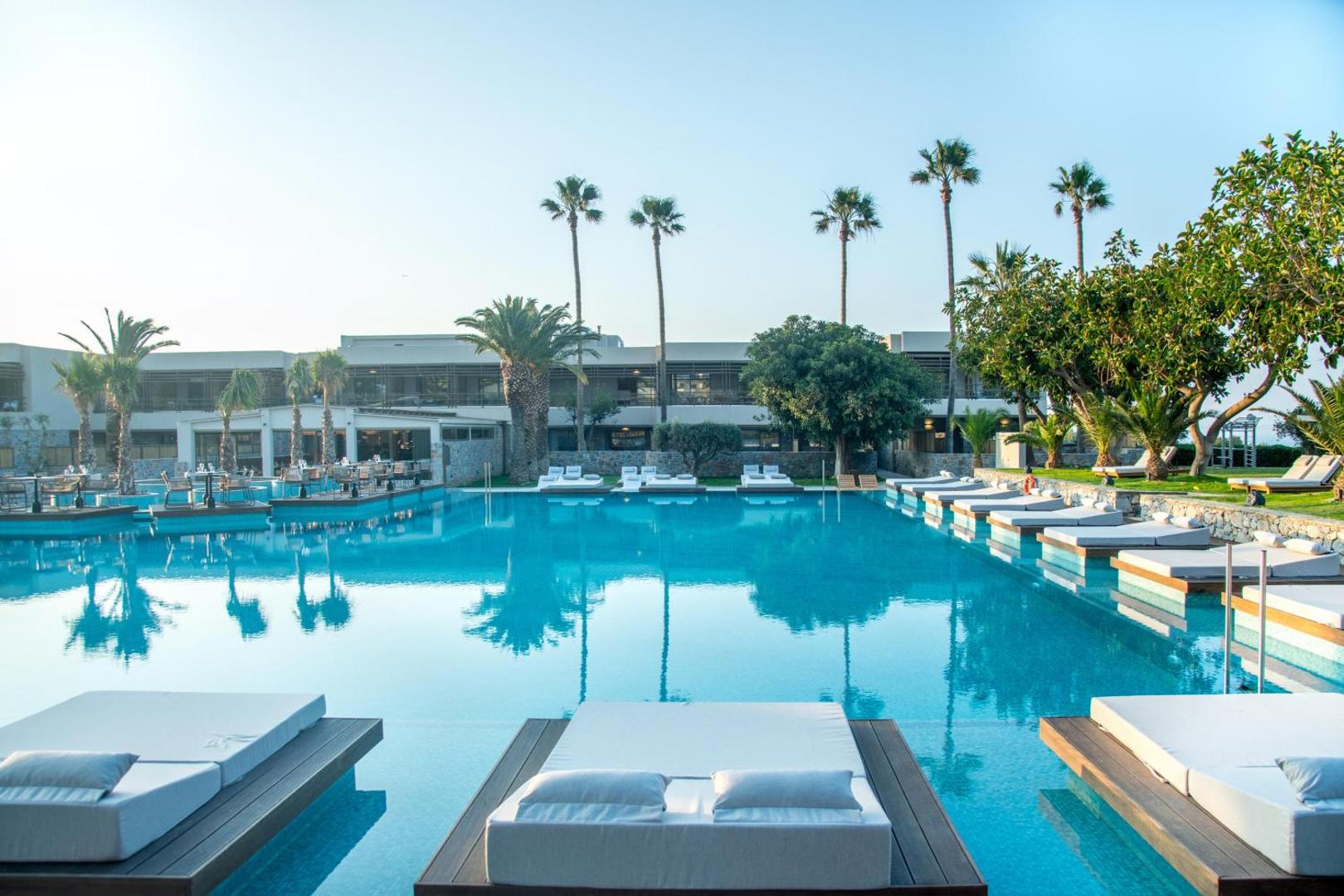 King Minos Retreat Resort & Spa galvenais āra baseins ar sauļošanās platformām ūdenī, palmām un viesnīcas ēku fonā Hersonisā