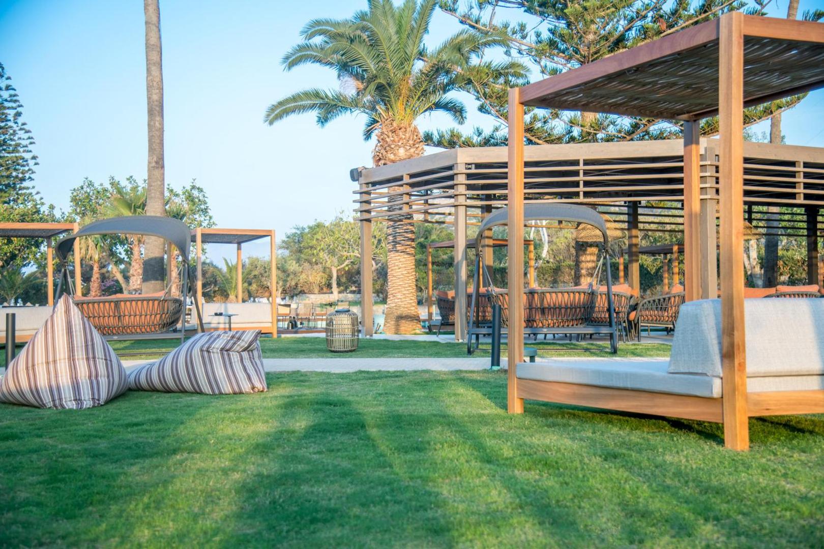 King Minos Retreat Resort & Spa dārza terase ar stilīgām nojumēm, šūpuļkrēsliem un palmām vakara saules gaismā