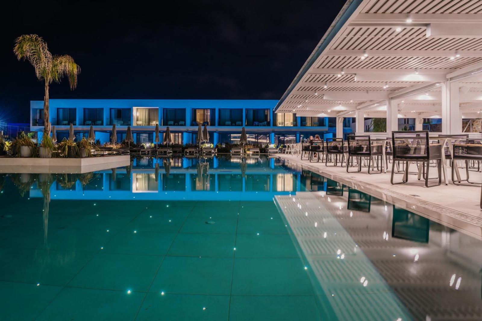 Gennadi Grand Resort izgaismots āra baseins naktī Rodas salā — moderna arhitektūra, terase ar galdiņiem un atspulgi ūdenī