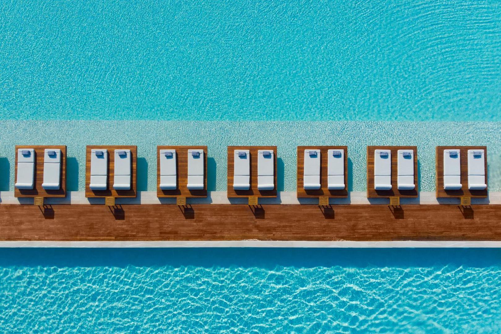 Gennadi Grand Resort baseina zona no augšas ar koka klāju, baltiem sauļošanās krēsliem un kristāldzidru ūdeni Rodas piekrastē