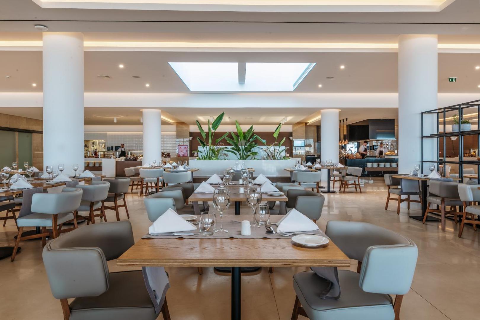 Gennadi Grand Resort galvenais restorāns ar modernu interjeru, gaišām kolonnām un klātiem galdiem — plaša un eleganta ēdināšanas zona