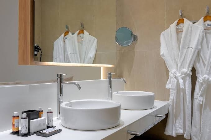 Gennadi Grand Resort vannasistabas interjers ar divām modernām izlietnēm, lielu spoguli un mīkstiem baltiem peldmēteļiem