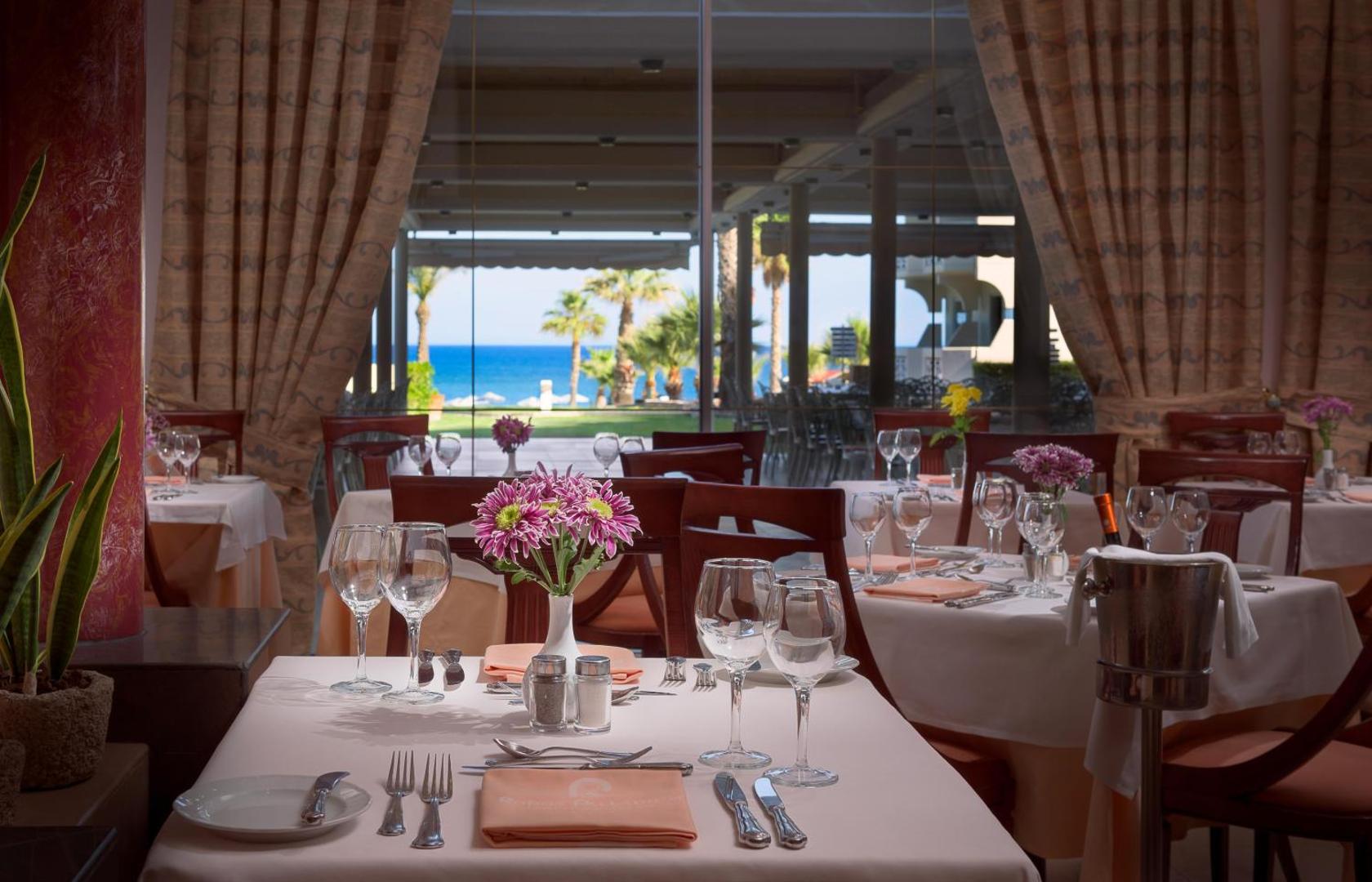 Rodos Palladium Leisure & Wellness iekštelpu restorāns ar servētiem galdiem un skatu caur logu uz dārzu
