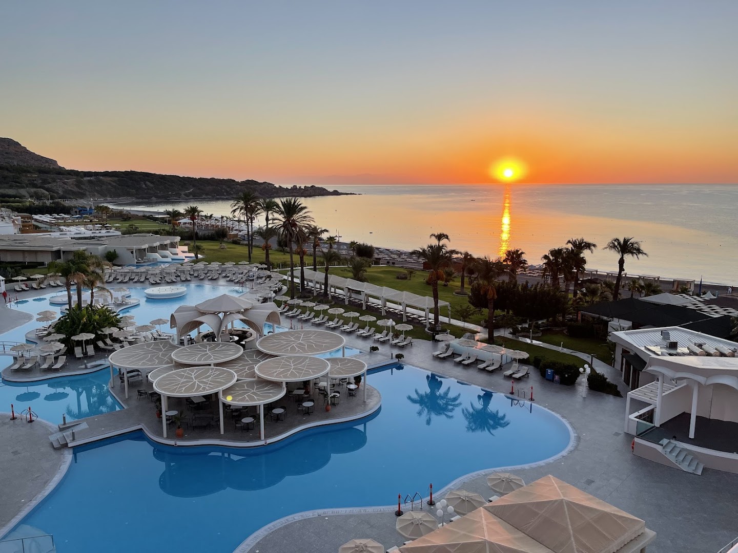 Saulriets Rodos Palladium Leisure & Wellness kūrortā ar skatu uz baseinu un Egejas jūru — panorāmas skats no viesnīcas terases vakarā