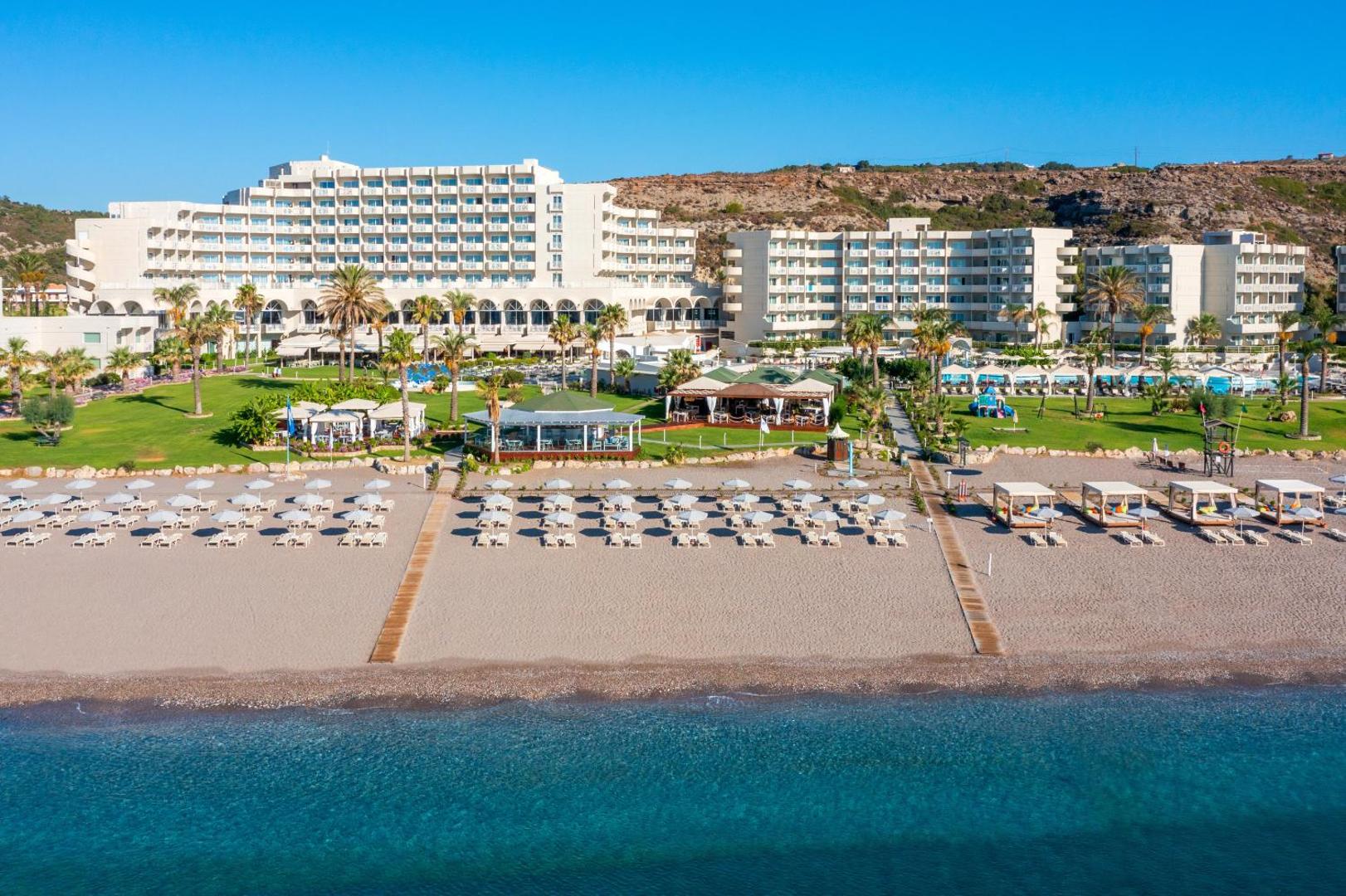 Rodos Palladium viesnīcas skats no jūras puses — labiekārtota pludmale ar saulessargiem, zālājs un galvenā ēka saulainā dienā