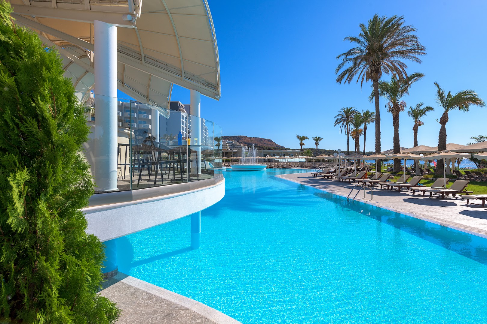 Rodos Palladium Leisure & Wellness plašais āra baseins ar palmām un sauļošanās krēsliem — fonā redzama strūklaka un zilās debesis Rodas piekrastē