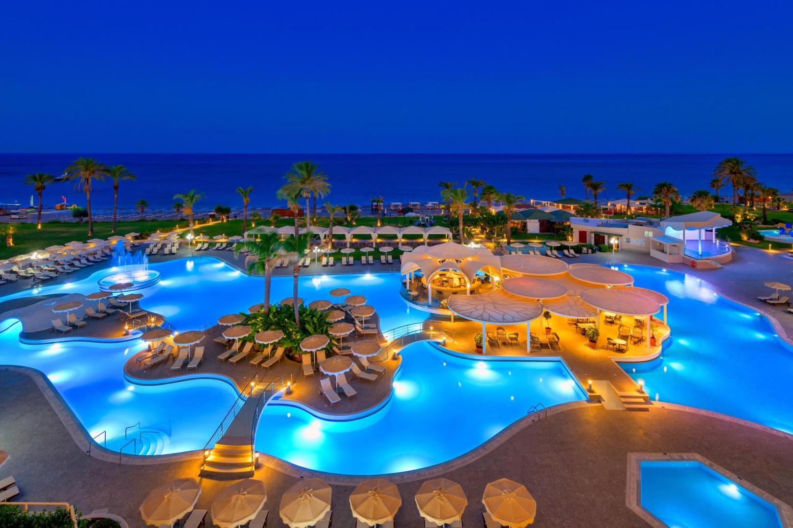 Rodos Palladium Leisure & Wellness iespaidīgs āra baseinu komplekss ar tiltiņiem un vakara apgaismojumu pašā jūras krastā Rodā.