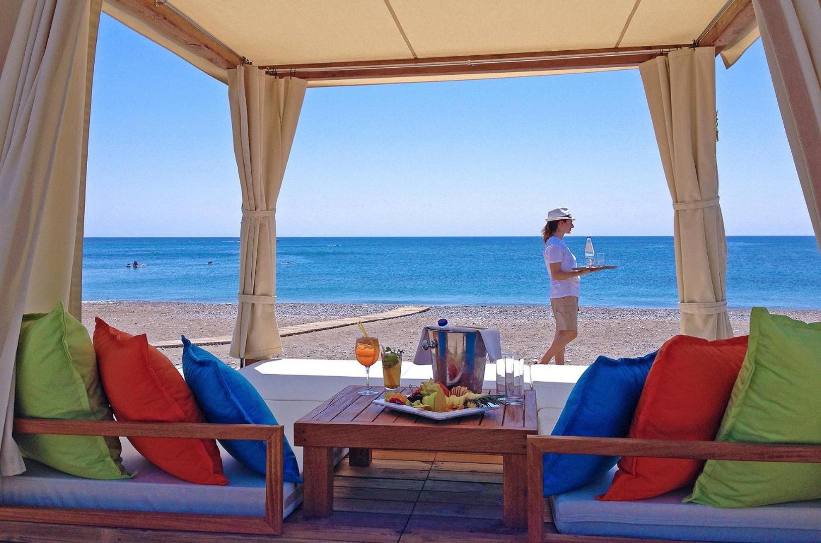 Atpūtas zona Rodos Palladium pludmales teltī ar krāsainiem spilveniem un uzkodām — skats uz zilo Vidusjūru