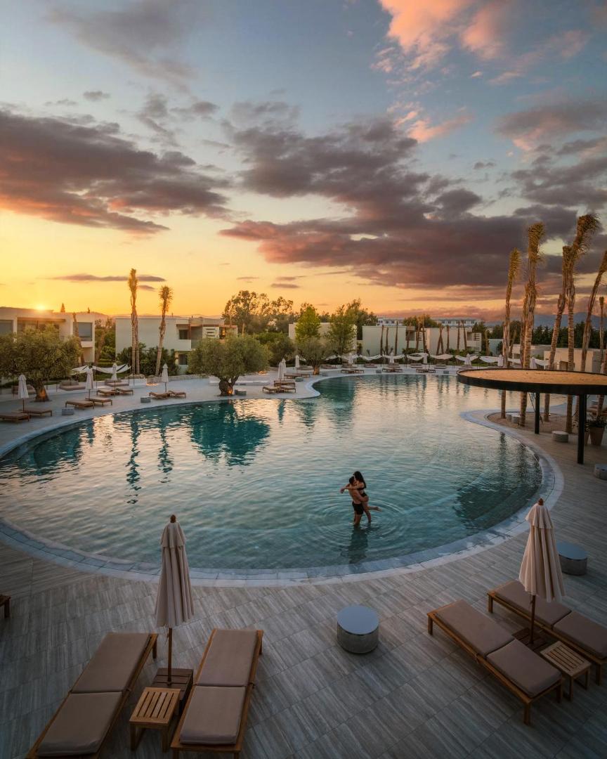 Sentido Port Royal Villas & Spa galvenais baseins saulrieta laikā Rodas salā ar atpūtas krēsliem un palmām fonā