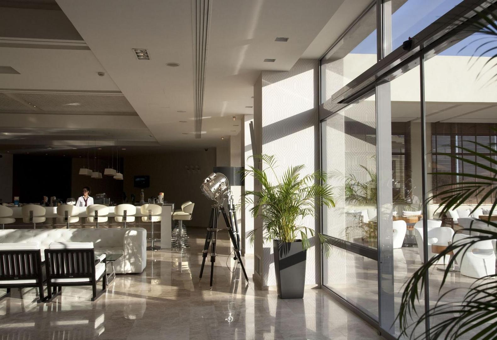 Sentido Port Royal Villas & Spa gaišais vestibila bārs ar lieliem logiem un modernām mēbelēm — skats uz saulaino atpūtas zonu