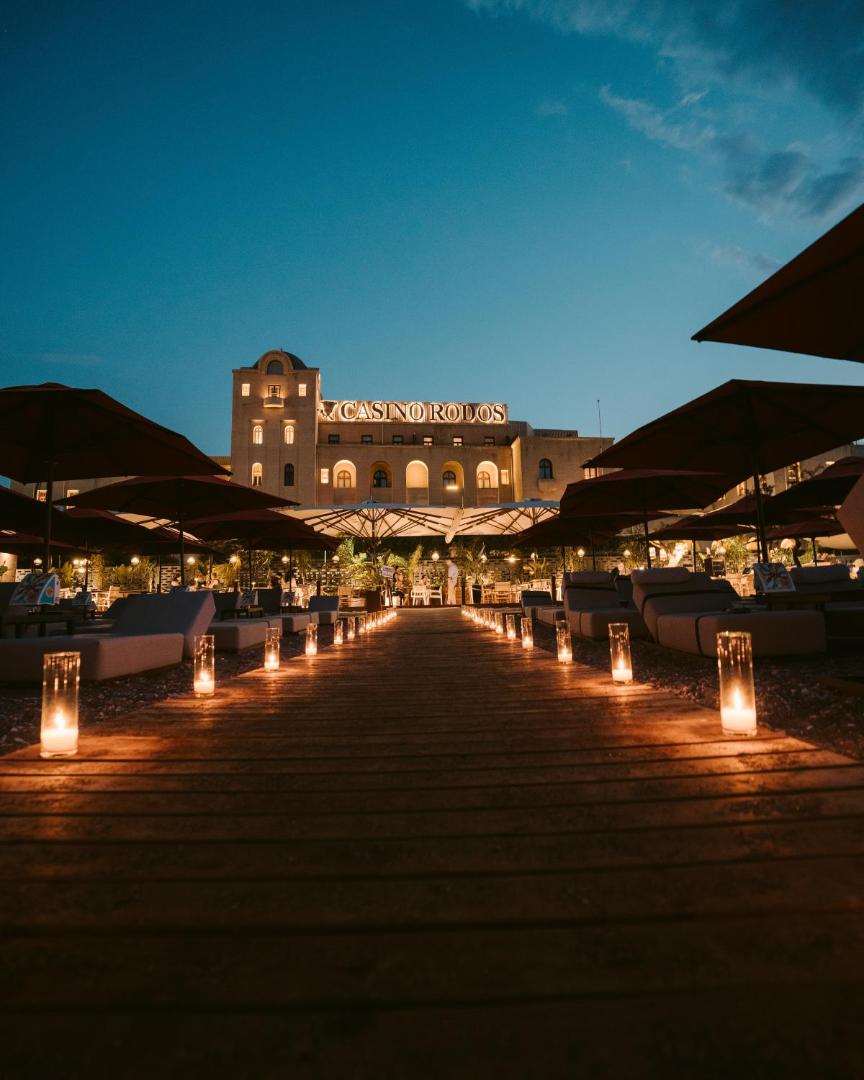 Vakara skats uz Grande Albergo Delle Rose vēsturisko ēku un Casino Rodos, izgaismota koka eja ar svecēm pludmales zonā.