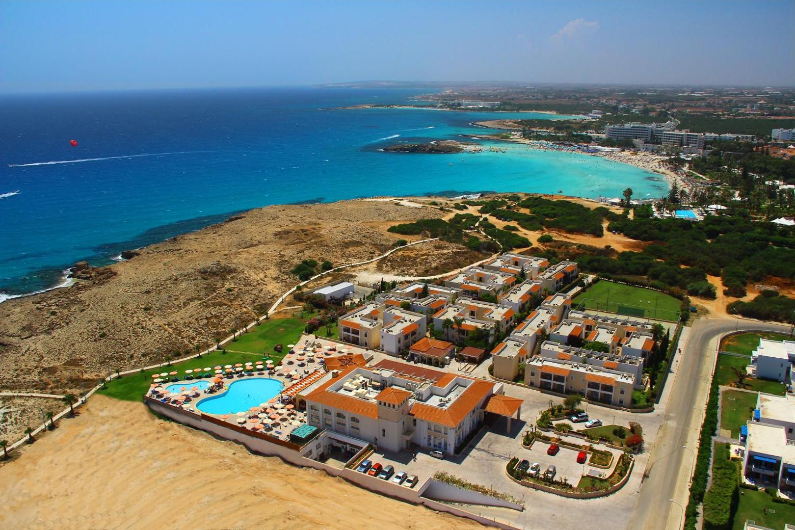 Aktea Beach Village viesnīcas panorāmas skats Ajanapā, Kiprā — komplekss ar āra baseinu, gaišām ēkām un tirkīzzilu jūras līci fonā