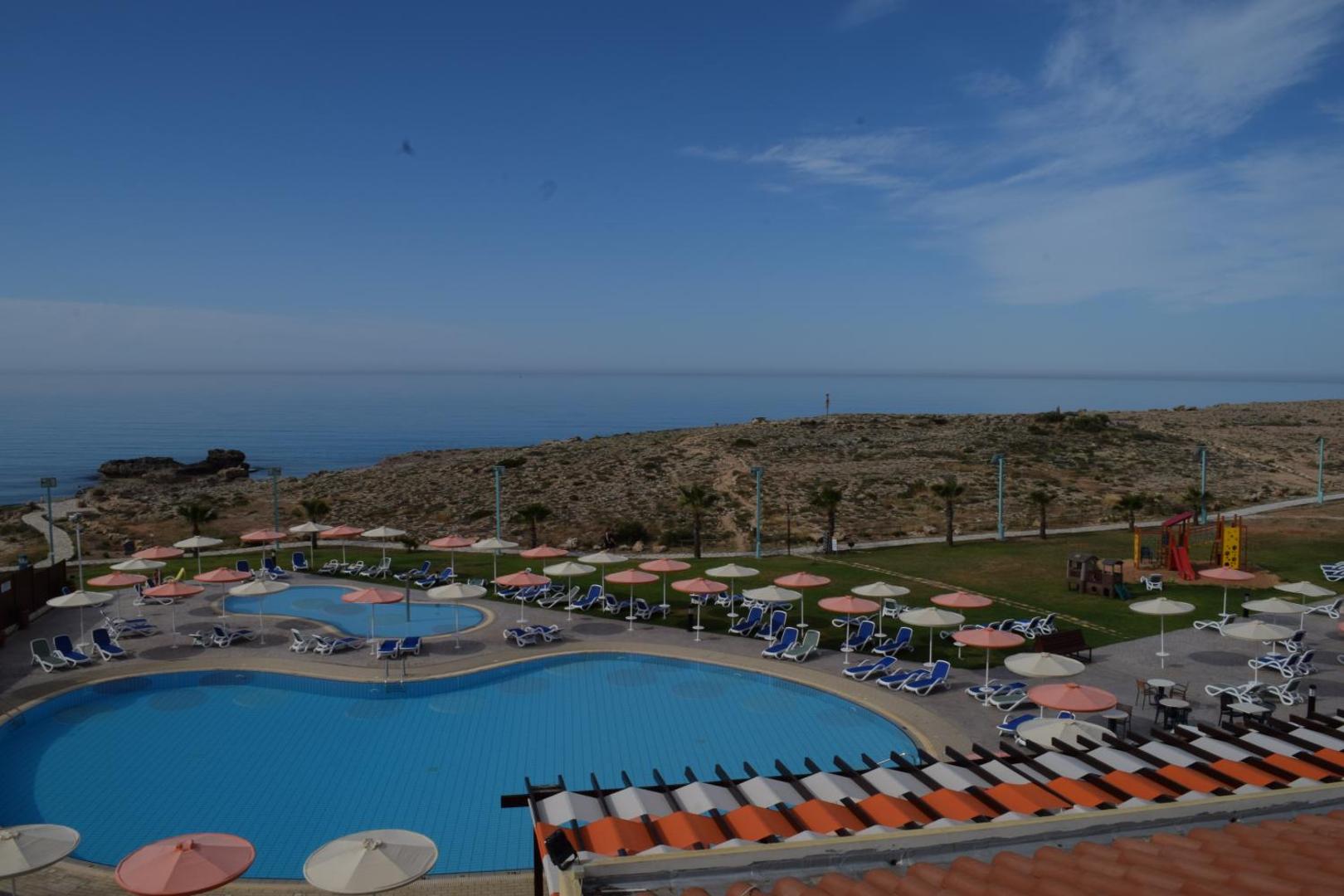 Aktea Beach Village āra baseinu zona ar saulessargiem un zviļņiem — skats uz Vidusjūras piekrasti un bērnu rotaļu laukumu