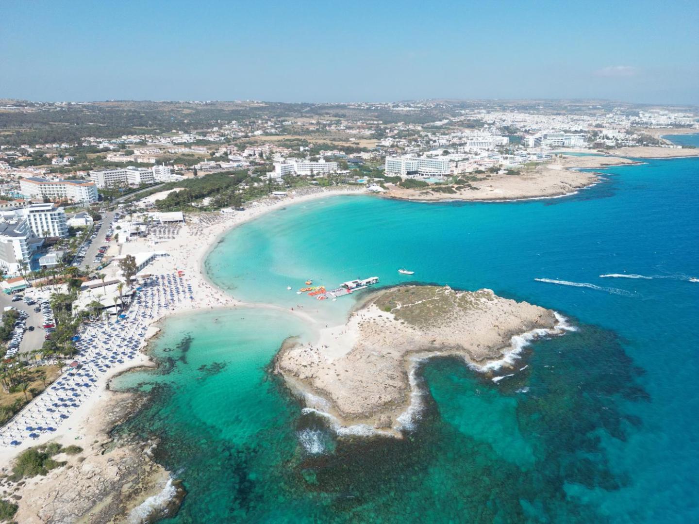 Aktea Beach Village skats no putna lidojuma uz Nissi pludmali Ajanapā, Kiprā — tirkīza ūdens, balto smilšu līcis un apkārtējās viesnīcas