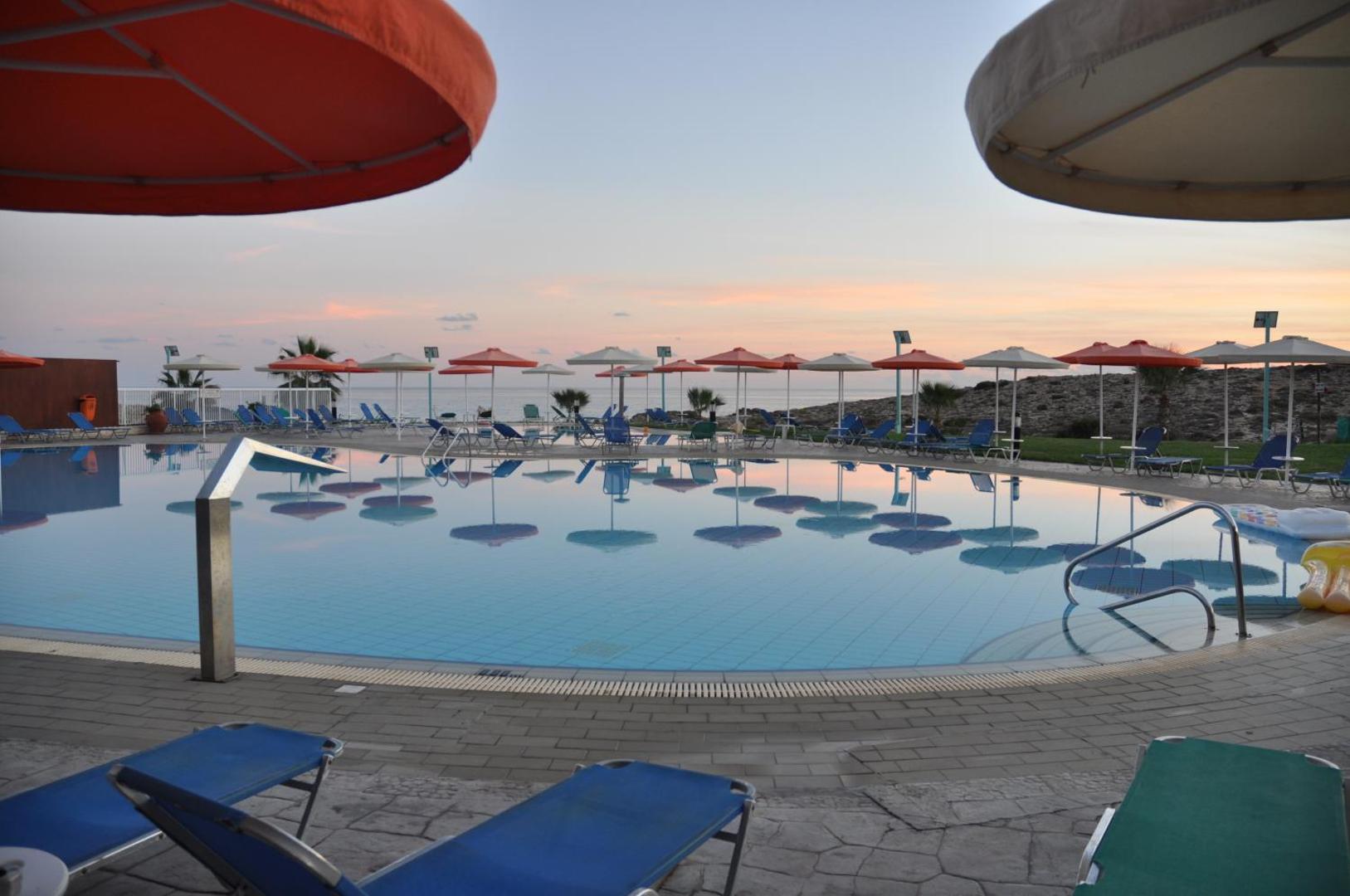 Aktea Beach Village āra baseins saulrieta laikā Aijanapā, Kiprā — atpūtas zona ar krāsainiem saulessargiem un zviļņiem pie ūdens malas