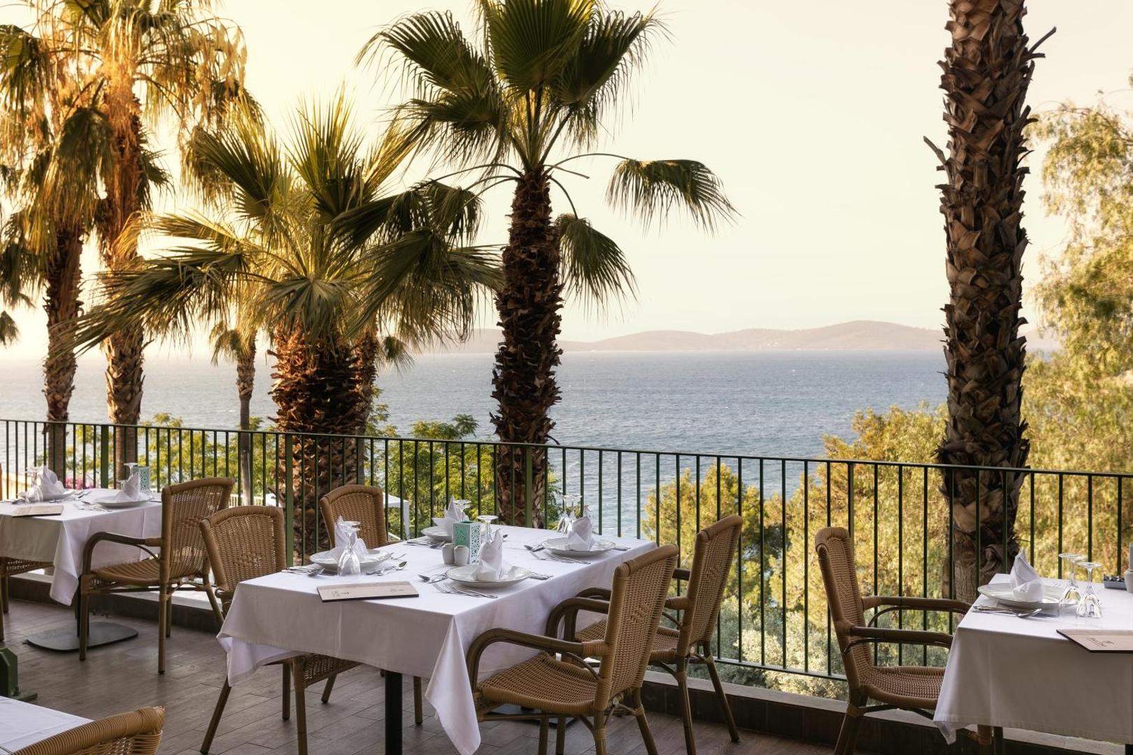 Viesnīcas Duja Bodrum restorāna terase ar galdiem, palmām un tiešu skatu uz Egejas jūru saulrieta gaismā