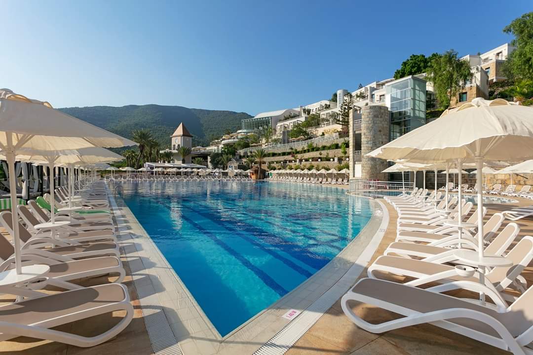 Plašs Duja Bodrum peldbaseins ar daudziem sauļošanās krēsliem un saulessargiem — fonā viesnīcas ēkas kalna nogāzē