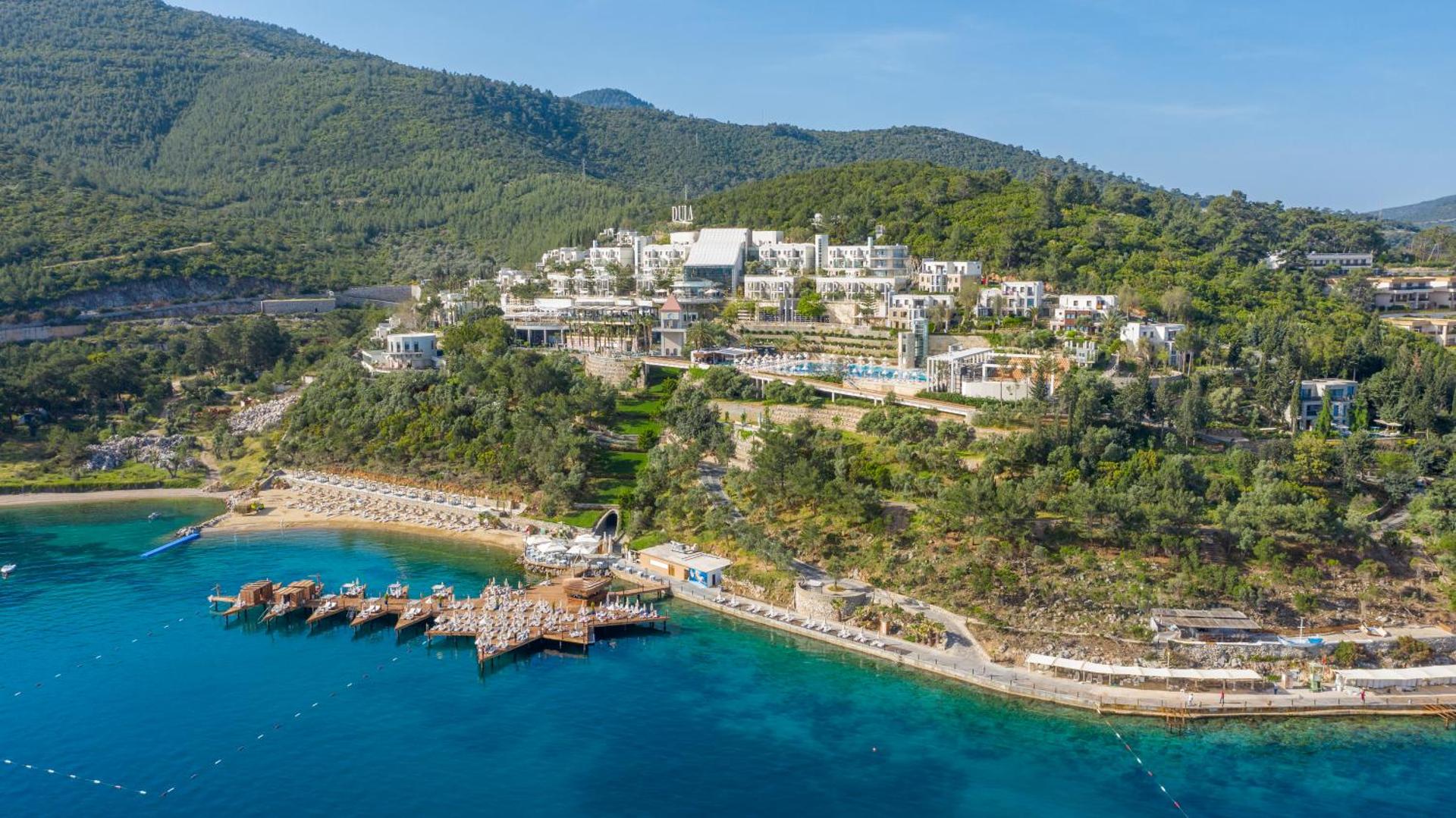 Duja Bodrum viesnīcas panorāmas skats no putna lidojuma — balta arhitektūra zaļā kalna nogāzē pie tirkīzzilas jūras ar privātu koka piestātni