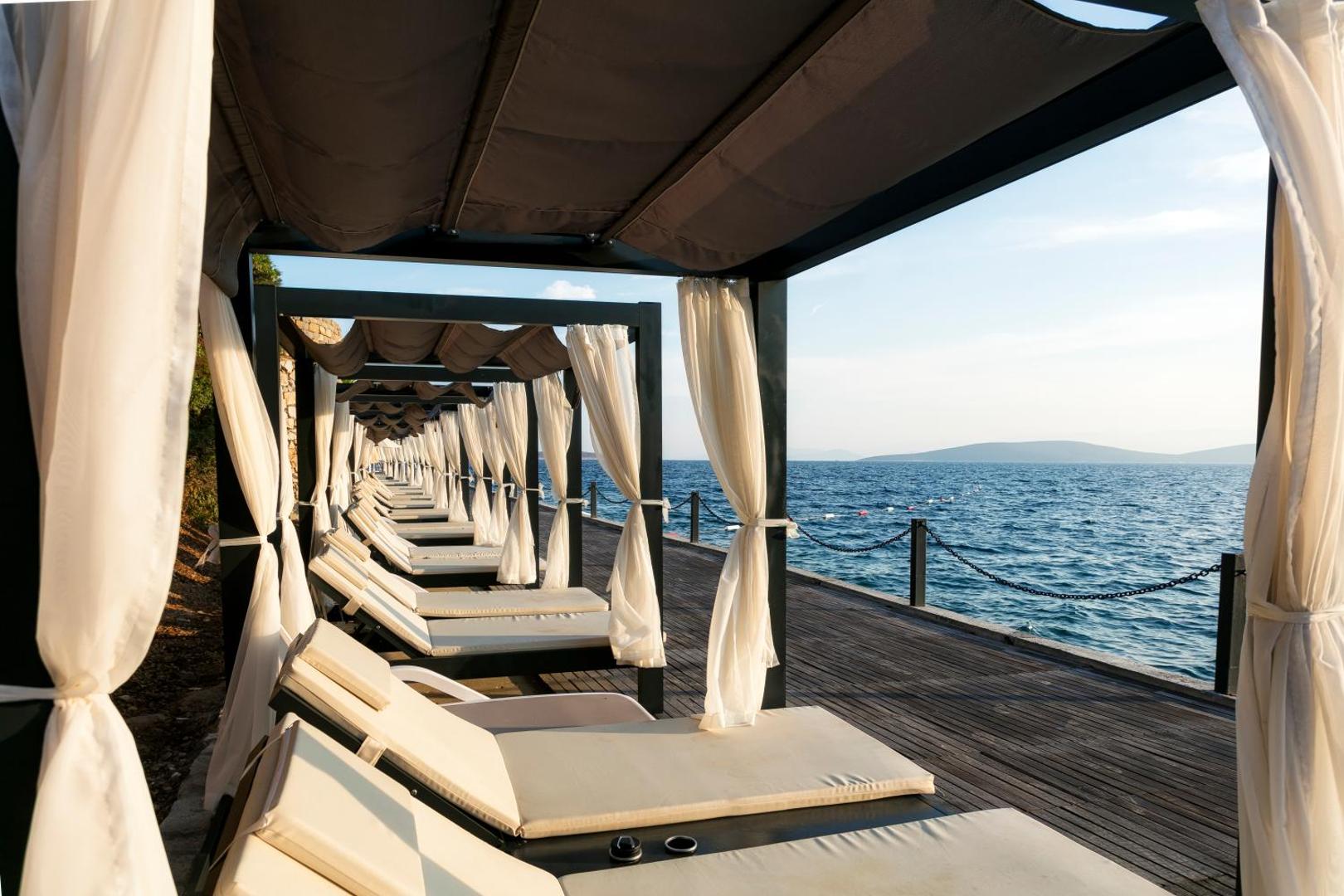 Ekskluzīvas atpūtas nojumes ar baltiem aizkariem un sauļošanās krēsliem uz koka klāja Duja Bodrum piekrastē
