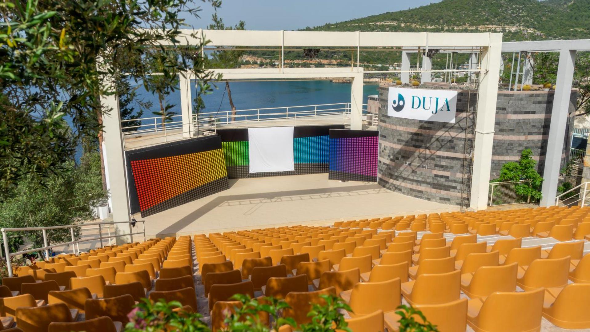 Duja Bodrum amfiteātris ar dzeltenām sēdvietām un skatuvi uz Egejas jūras un kalnu fona Bodrumas piekrastē