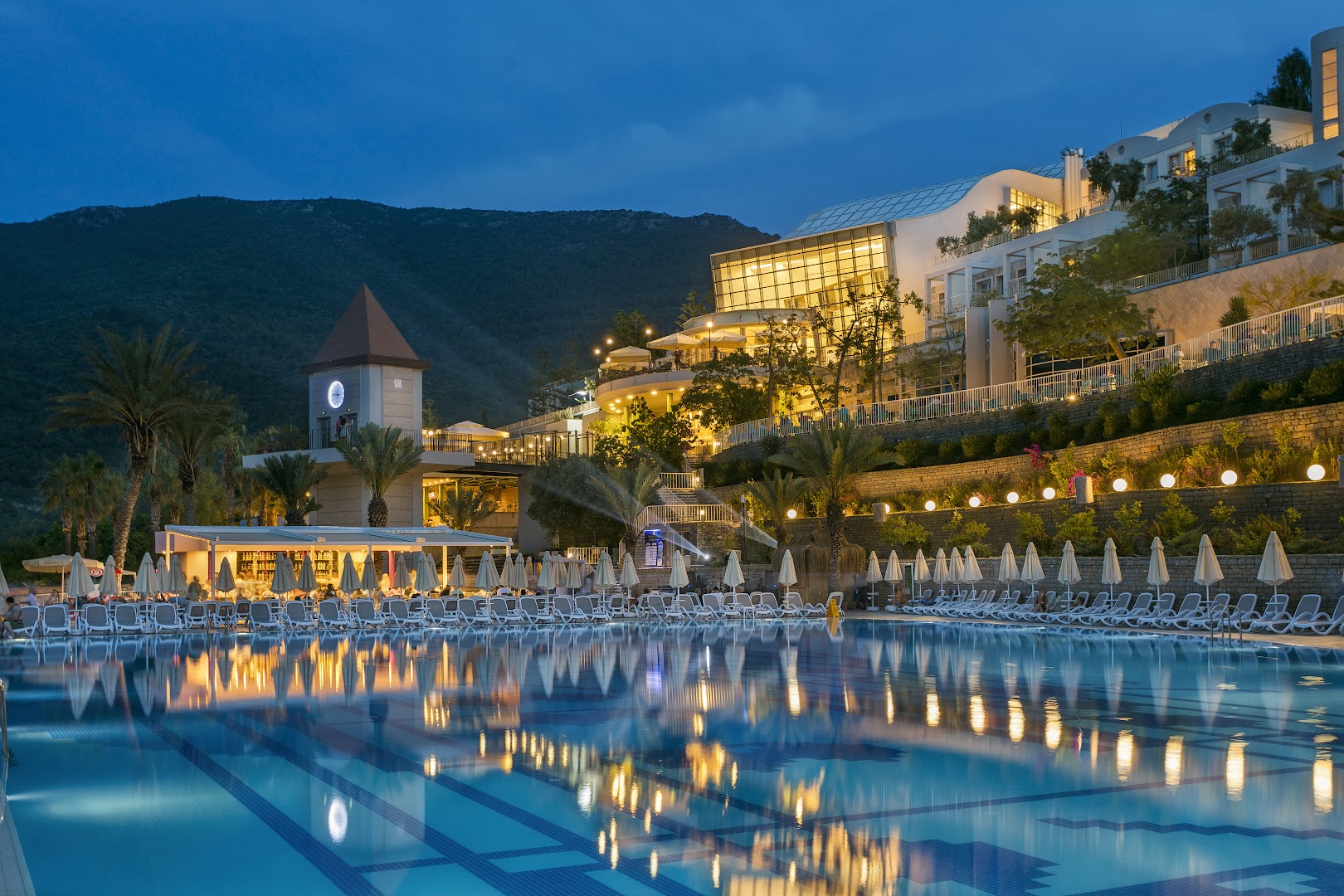 Duja Bodrum viesnīcas teritorija vakarā — izgaismots baseins, restorāna terase un kalnu siluets saulrietā