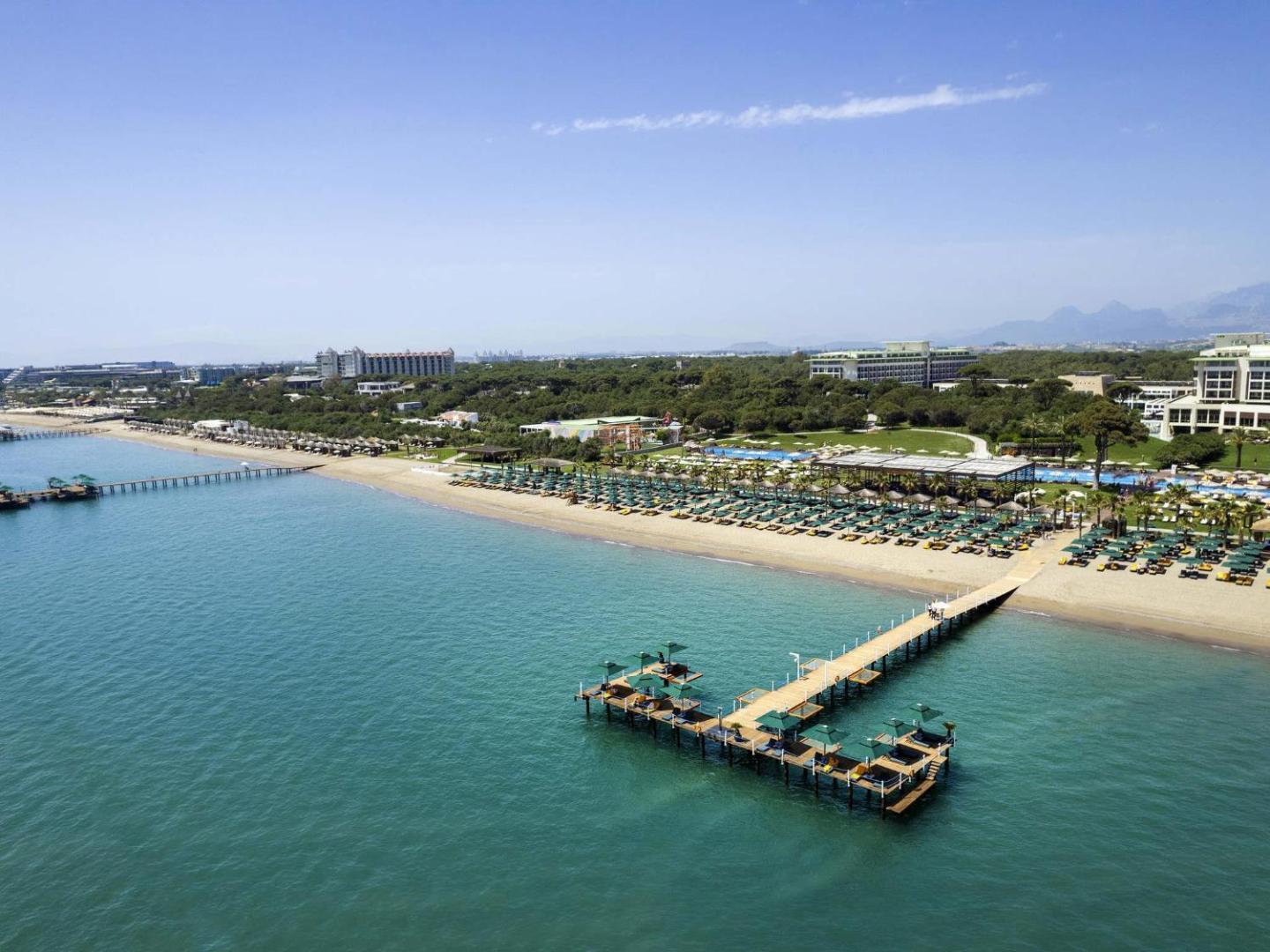 Rixos Premium Belek panorāmas skats no putna lidojuma uz plašo pludmali, koka piestātnēm un kūrorta teritoriju pie Vidusjūras