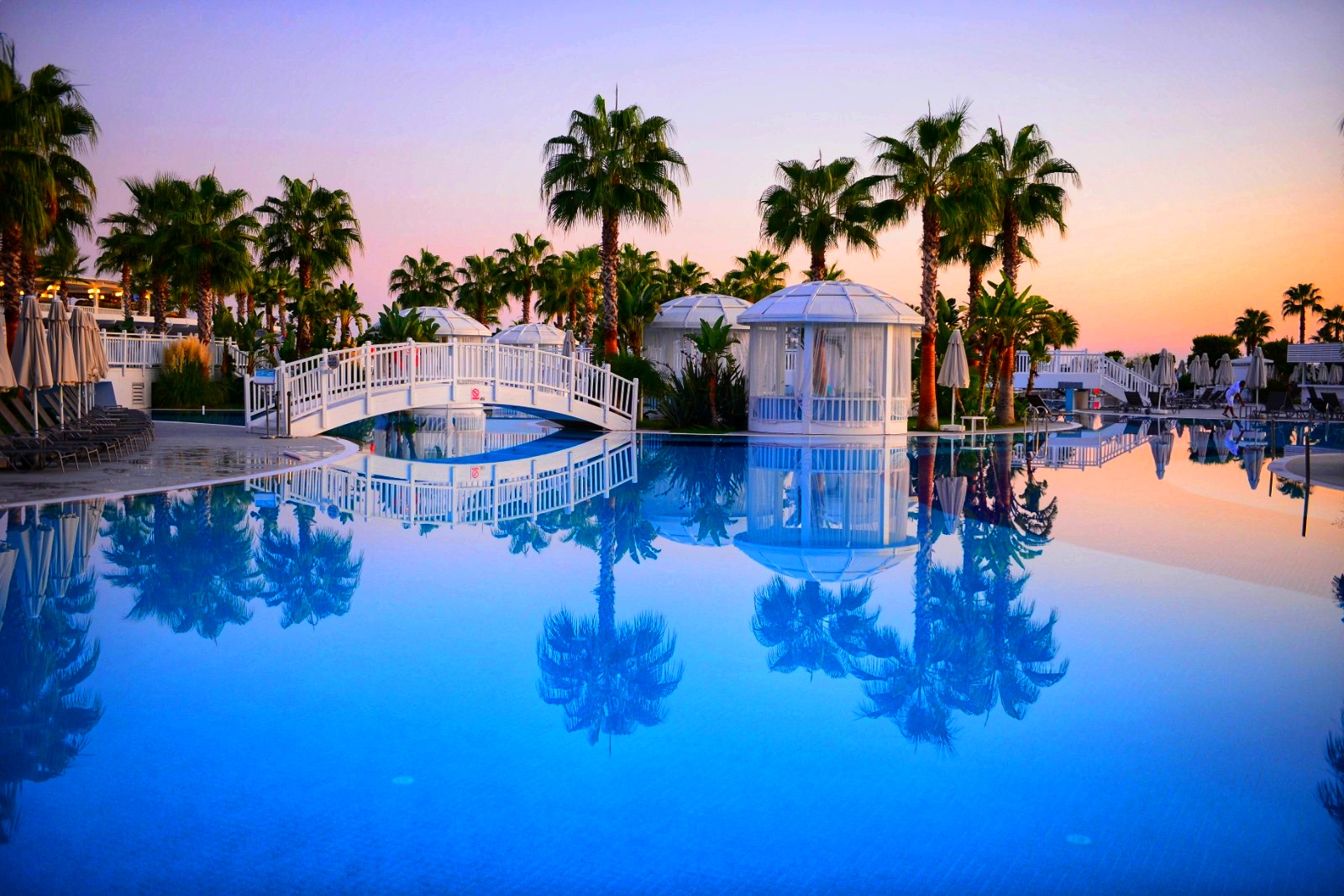 Sueno Hotels Deluxe Belek baseins krēslas stundā ar palmu atspulgu ūdenī, dekoratīvu tiltiņu un baltām atpūtas lapenēm