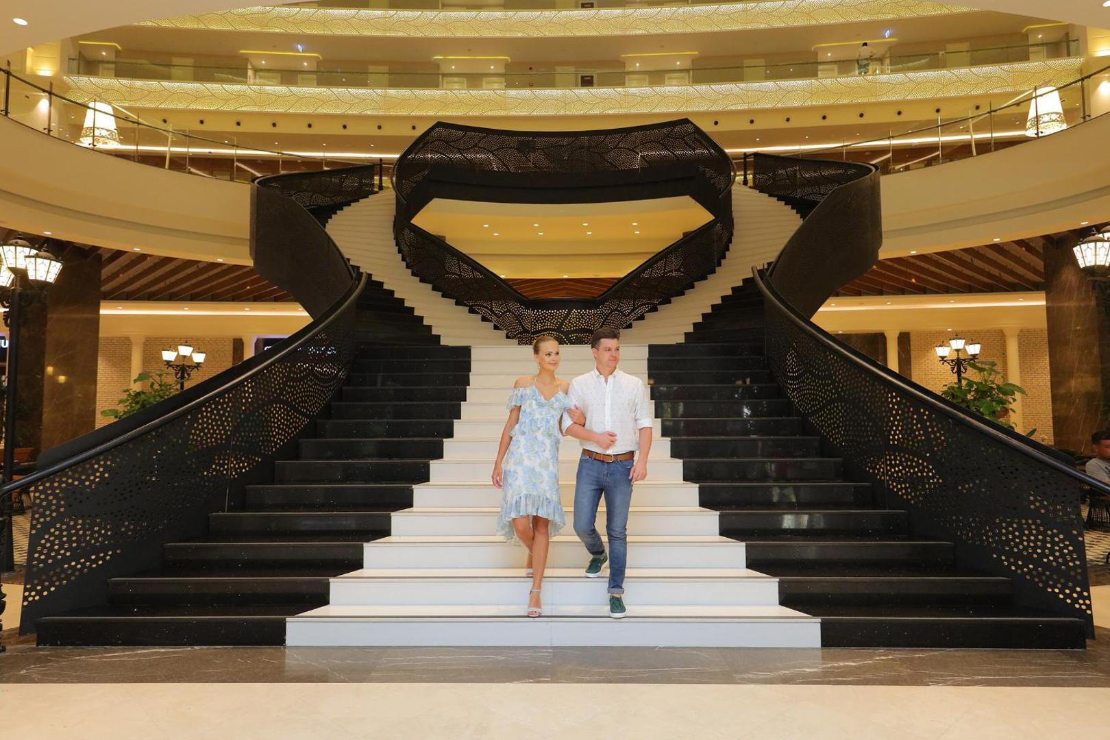 Sueno Hotels Deluxe Belek greznas kāpnes viesnīcas vestibilā ar pāri, kas dodas lejup pa melna marmora pakāpieniem