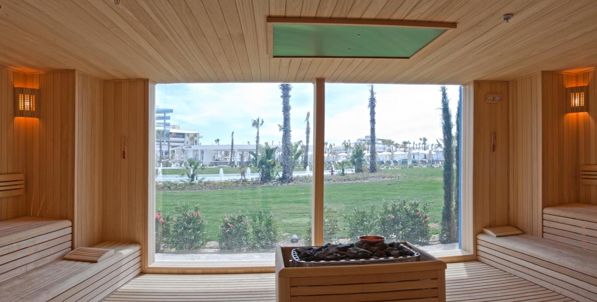 Sueno Hotels Deluxe Belek spa centra sauna ar lielu panorāmas logu uz viesnīcas dārzu un palmām Belekā, Turcijā