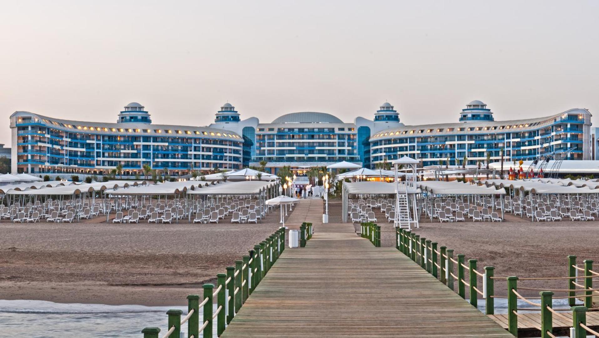 Sueno Hotels Deluxe Belek fasāde un privātā pludmale ar koka laipu — saulrieta gaisma virs sauļošanās krēsliem un nojumēm
