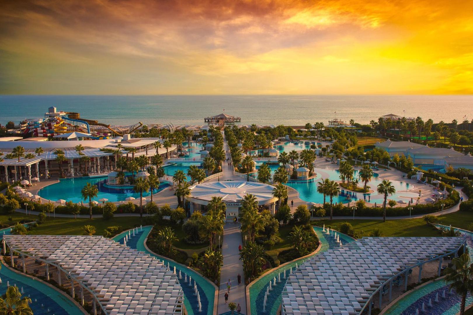 Sueno Hotels Deluxe Belek panorāmas skats saulrietā uz viesnīcas teritoriju, baseiniem un Vidusjūras piekrasti Antaljas reģionā