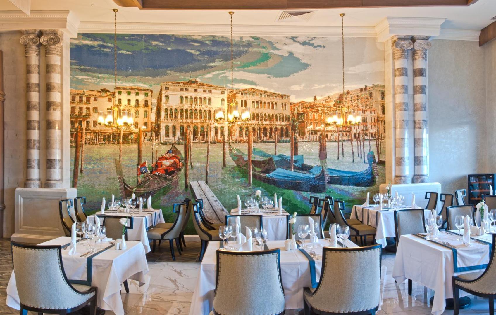 Sueno Hotels Deluxe Belek restorāna zāle ar lielu mozaīku pie sienas, kurā attēlota Venēcija — balti klāti galdi