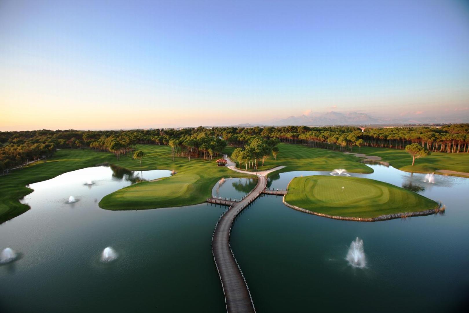 Sueno Hotels Deluxe Belek golfa laukumu panorāmas skats ar ezeriem, tiltiņiem un priežu mežu Belekā saulrietā