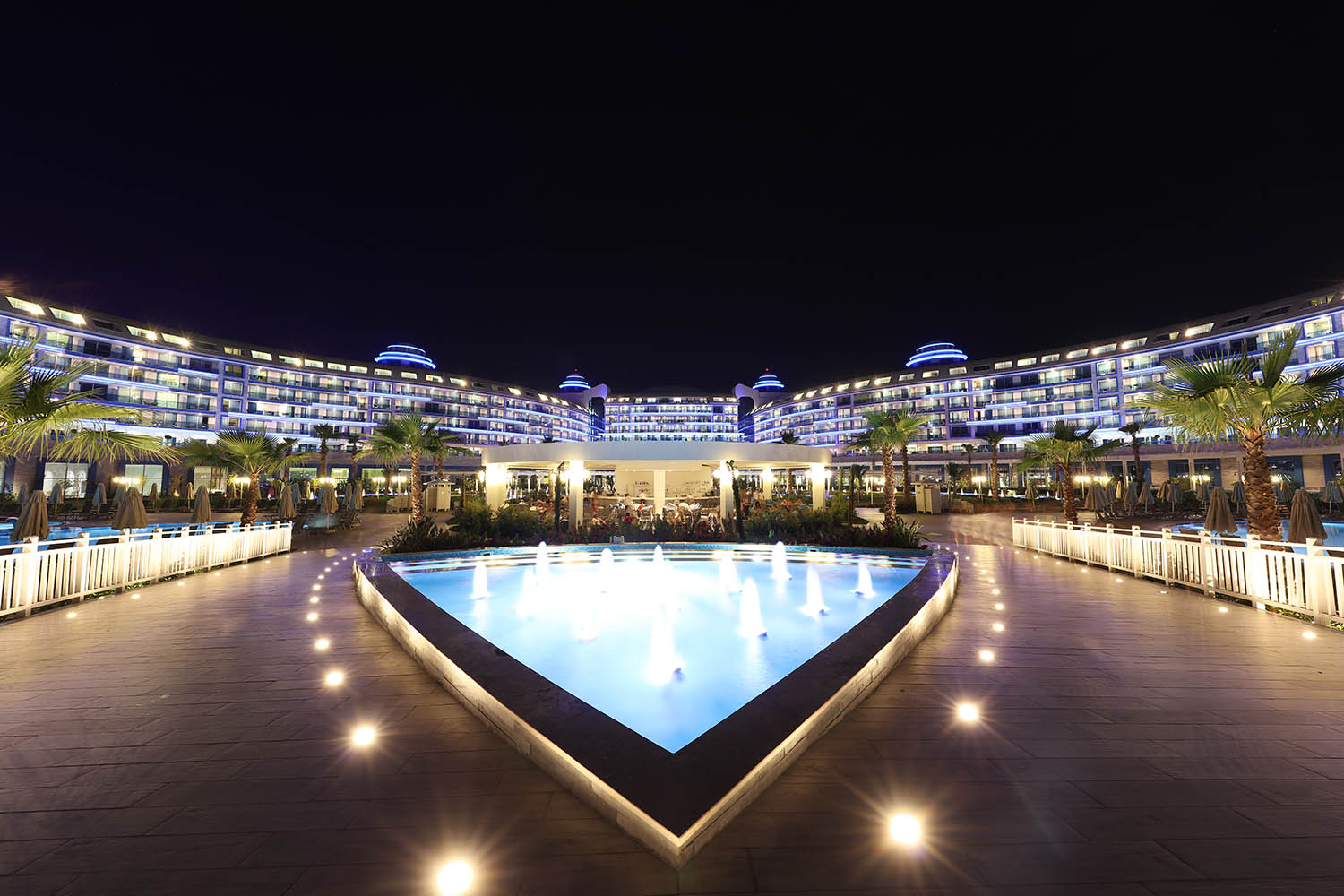 Sueno Hotels Deluxe Belek viesnīcas pagalms naktī ar izgaismotu strūklaku, palmām un modernu fasādes apgaismojumu