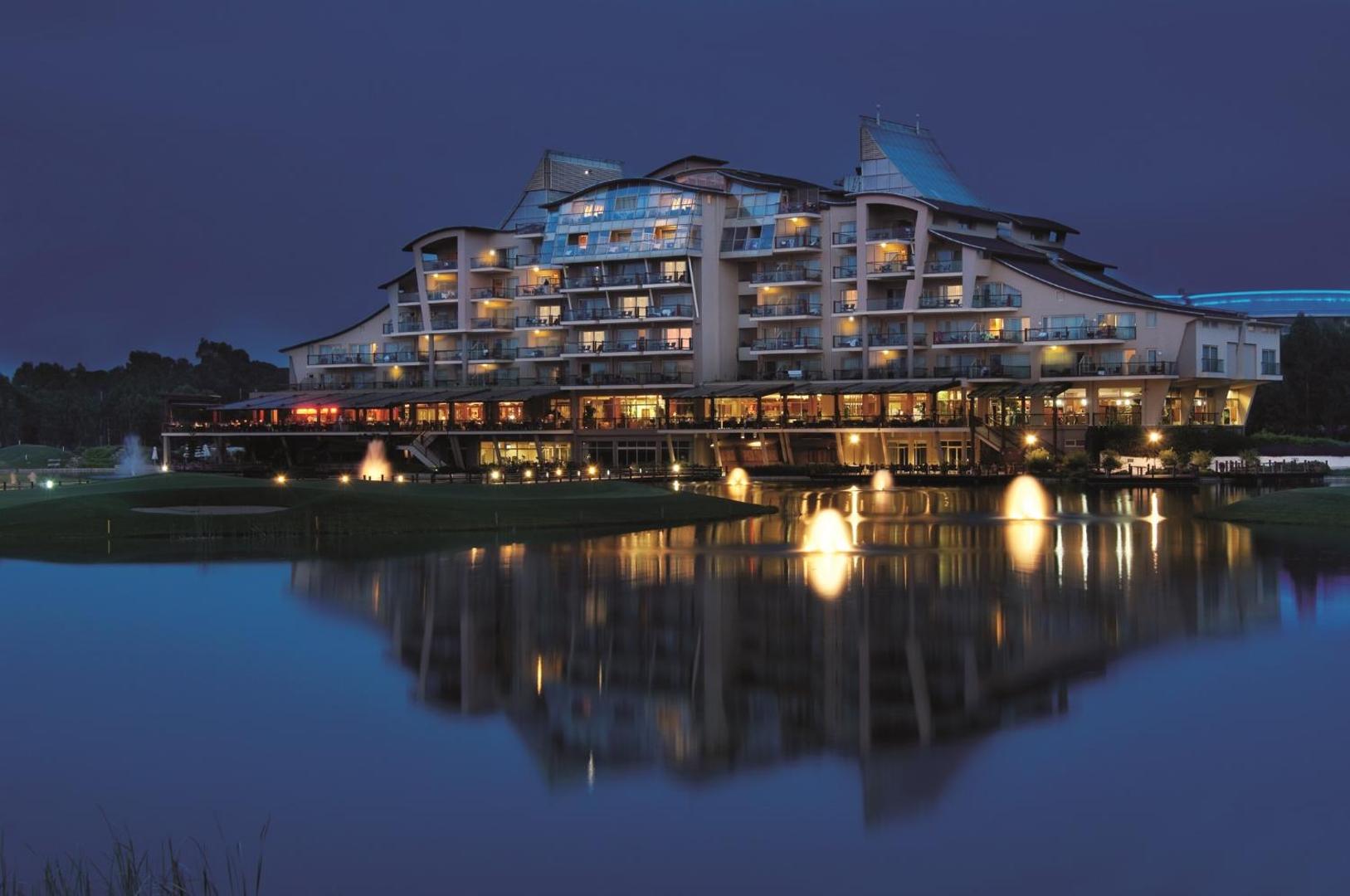 Sueno Hotels Golf Belek viesnīcas fasāde vakarā ar izgaismotām strūklakām ezerā un atspulgu ūdenī — romantiska atmosfēra Belekā