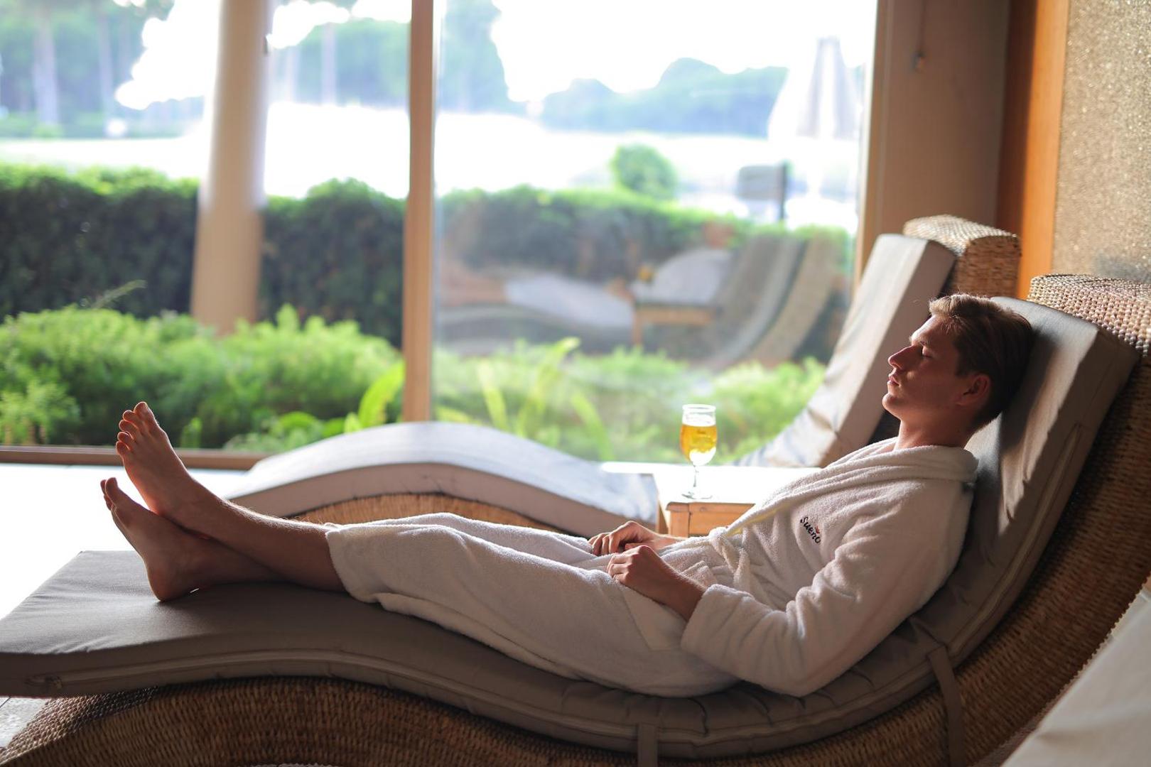 Viesis baltā peldmētelī atpūšas uz zvilņa Sueno Hotels Golf Belek spa relaksācijas zonā ar skatu uz dārzu