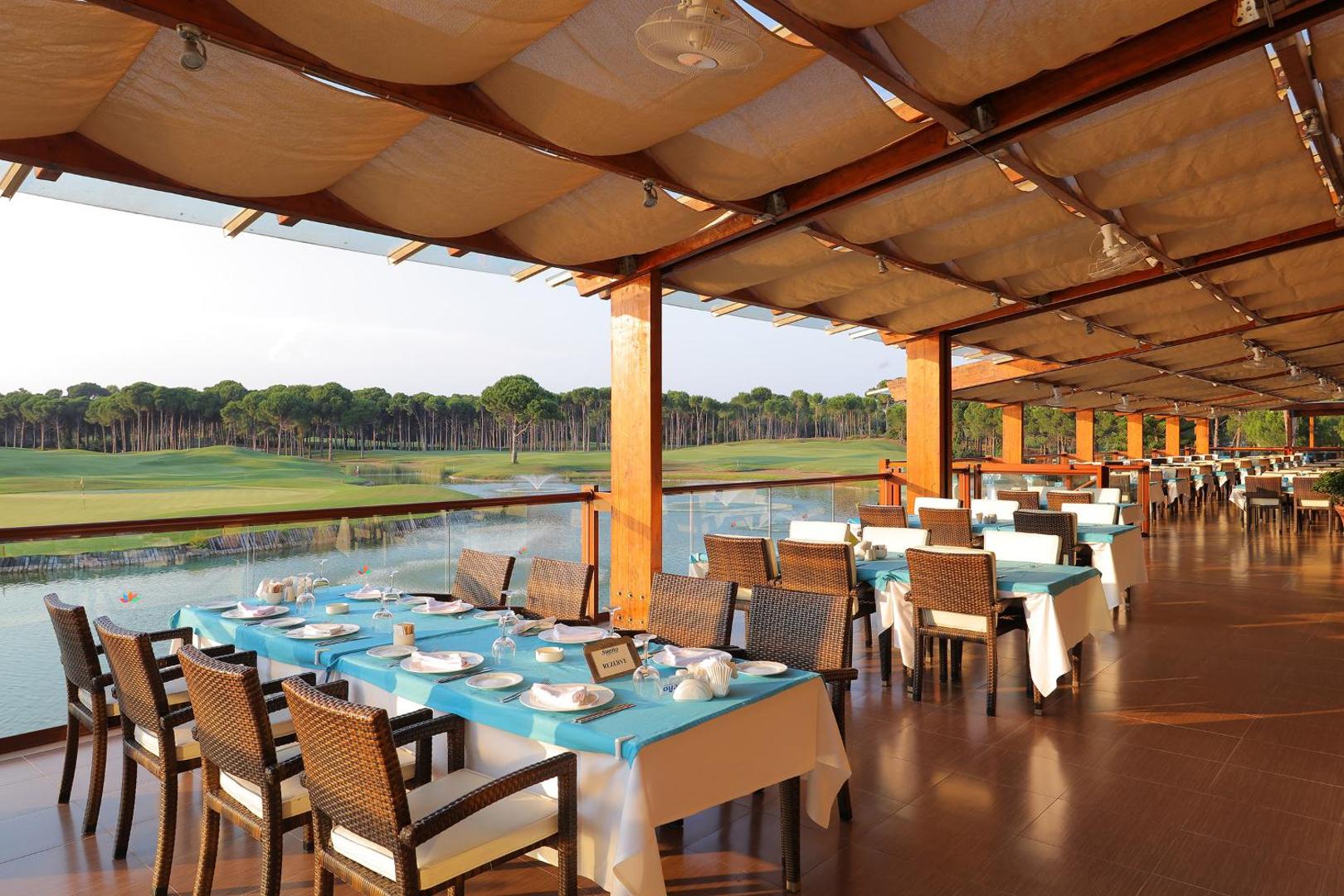 Sueno Hotels Golf Belek restorāna terase ar skatu uz golfa laukumiem un ezeru Belekā, Turcijā — klāti galdi ar gaiši ziliem galdautiem