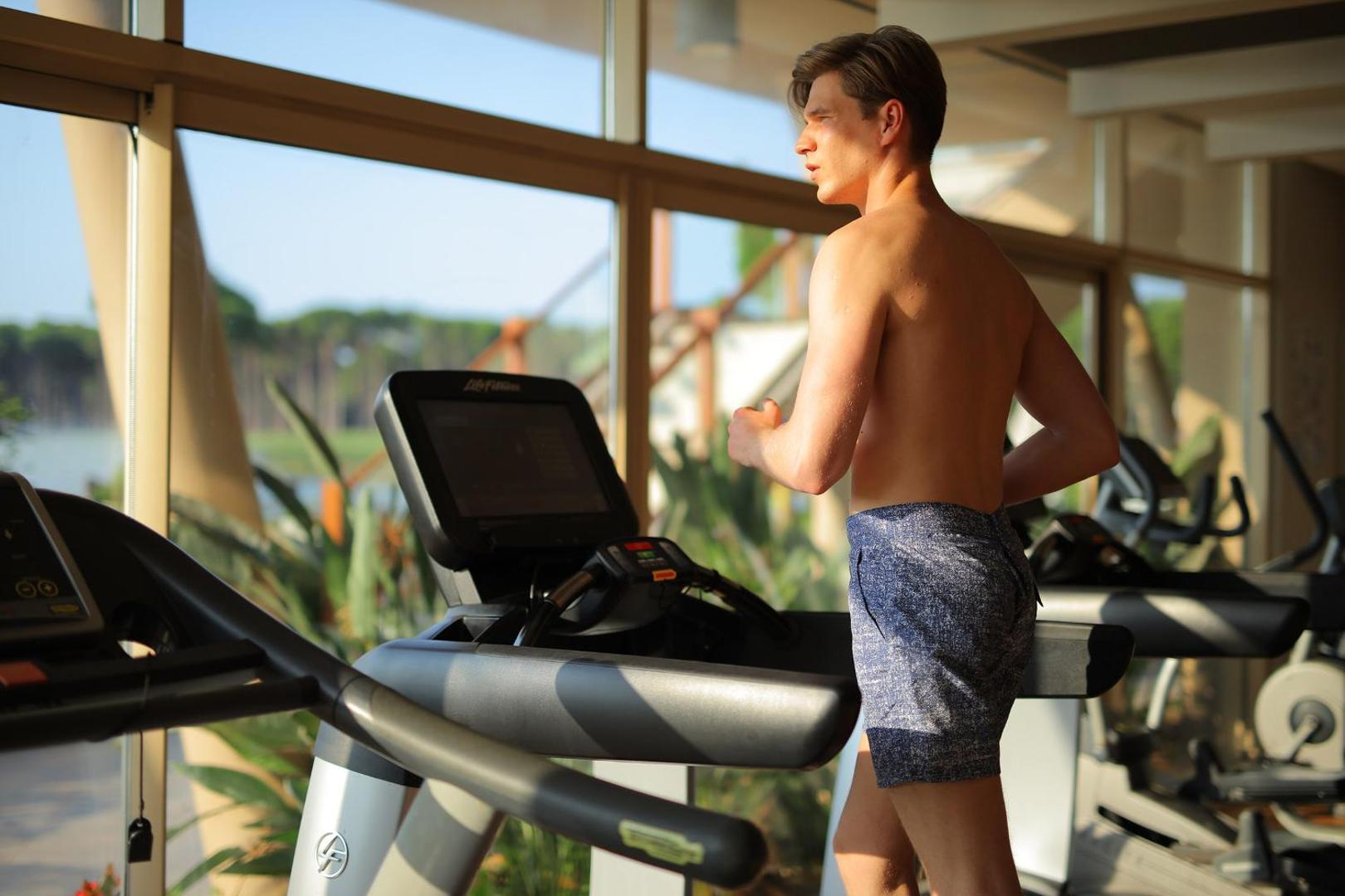 Vīrietis sporto Sueno Hotels Golf Belek fitnesa centrā uz skrejceliņa ar skatu caur logu uz dabu