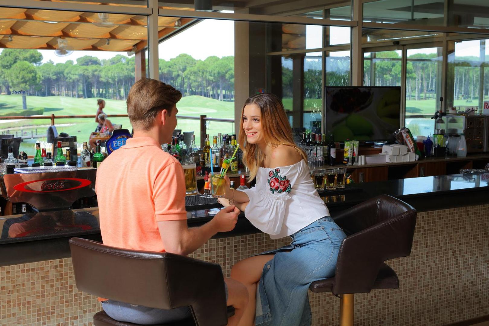 Pāris bauda kokteiļus Sueno Hotels Golf Belek bārā ar skatu uz plašajiem golfa laukumiem un priežu mežu