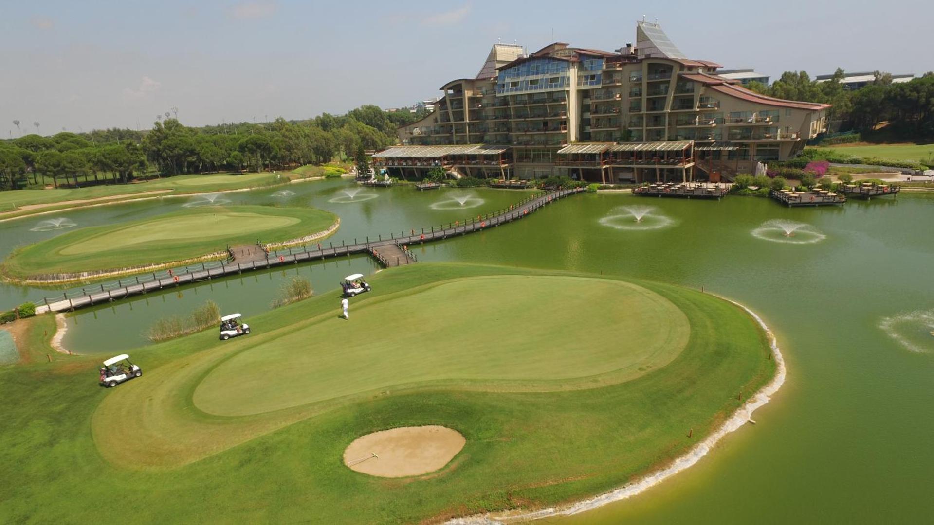 Sueno Hotels Golf Belek golfa laukums ar ezeru un strūklakām — fonā redzama viesnīcas ēka un golfa mašīnas uz zaļā zālāja
