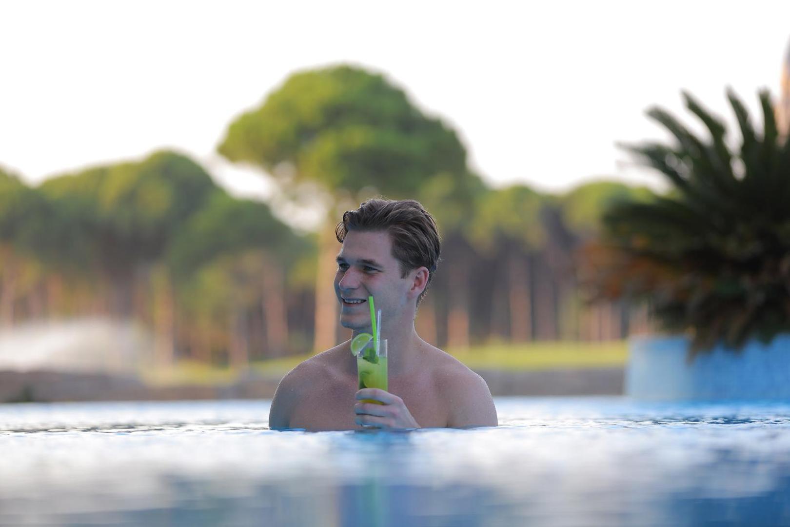 Vīrietis bauda kokteili Sueno Hotels Golf Belek āra baseinā uz zaļojošu priežu un dabas ainavas fona saulrietā