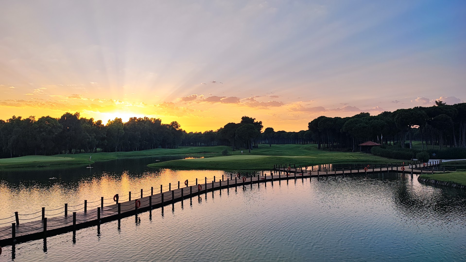 Saulriets virs Sueno Hotels Golf Belek golfa laukumiem un ezera ar koka gājēju tiltu — mierpilna vakara ainava Belekā