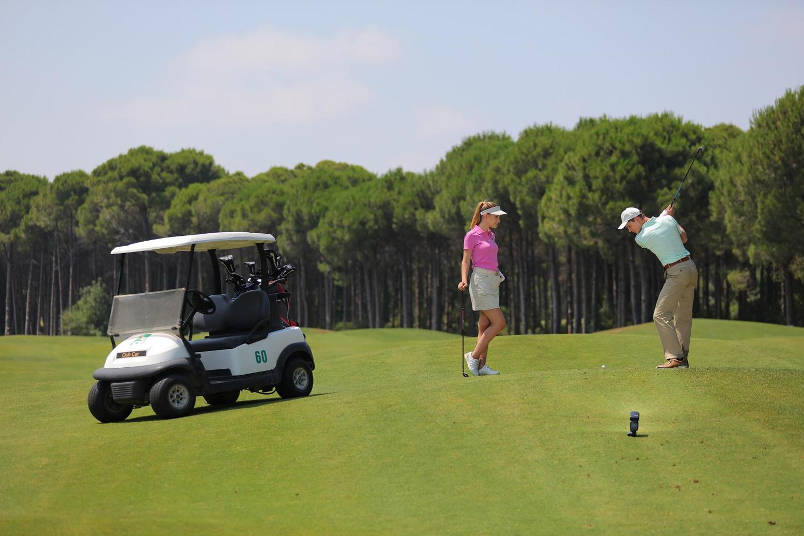 Viesi spēlē golfu Sueno Hotels Golf Belek kūrortā, fonā golfa automašīna un augstas priedes saulainā dienā