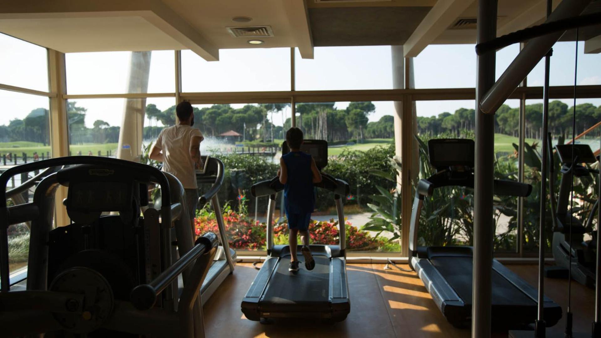 Sueno Hotels Golf Belek trenažieru zāle ar panorāmas logiem, no kuriem paveras skats uz golfa laukumiem un strūklaku