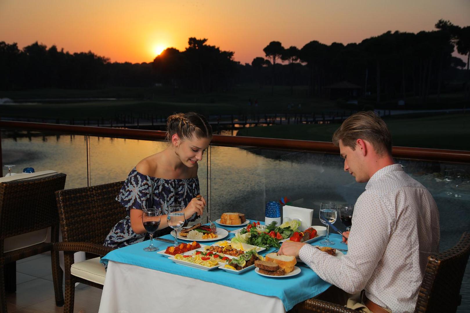 Pāris bauda vakariņas saulrietā uz Sueno Hotels Golf Belek terases ar skatu uz ezeru un priežu mežu Belekas reģionā