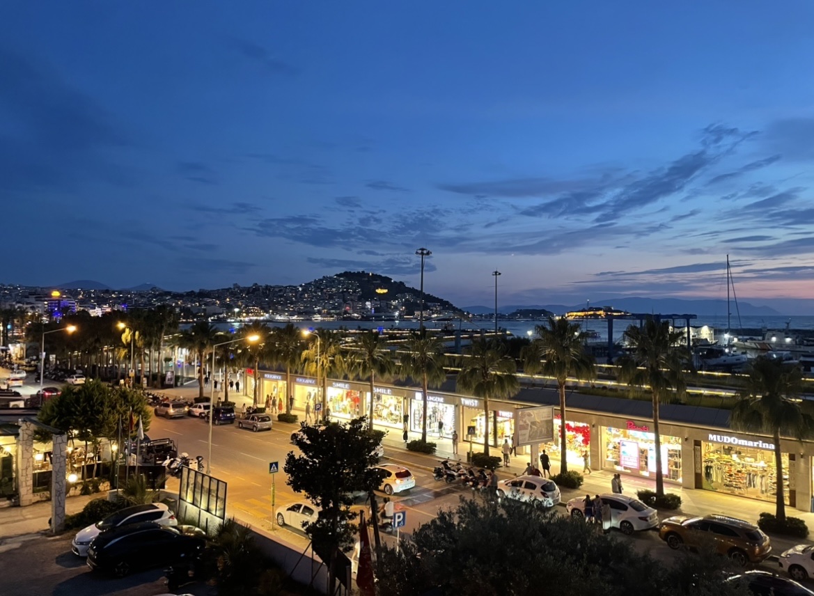 Paloma Marina Suites apkārtne vakarā ar izgaismotu pilsētas ielu, veikaliem, palmām un Kusadasi pilsētas panorāmu fonā